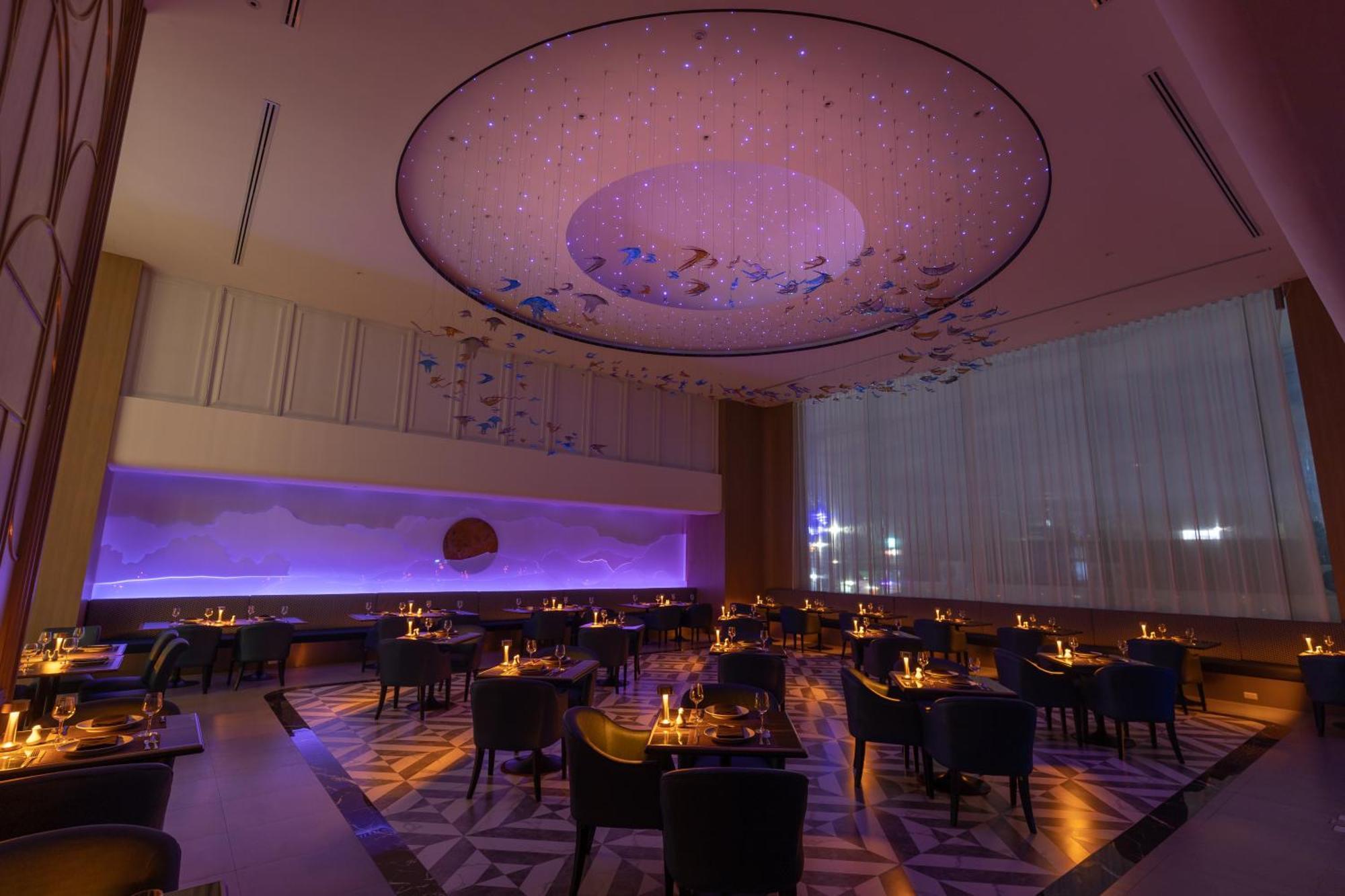 Siam Mandarina Hotel - Free Bkk Airport Shuttle Samut Prakan Ngoại thất bức ảnh The photo shows a modern and elegant restaurant interior. The ceiling is adorned with a striking circular chandelier, which features a soft purple hue and is surrounded by artistic elements resembling fish or marine life. Below, the dining area is se